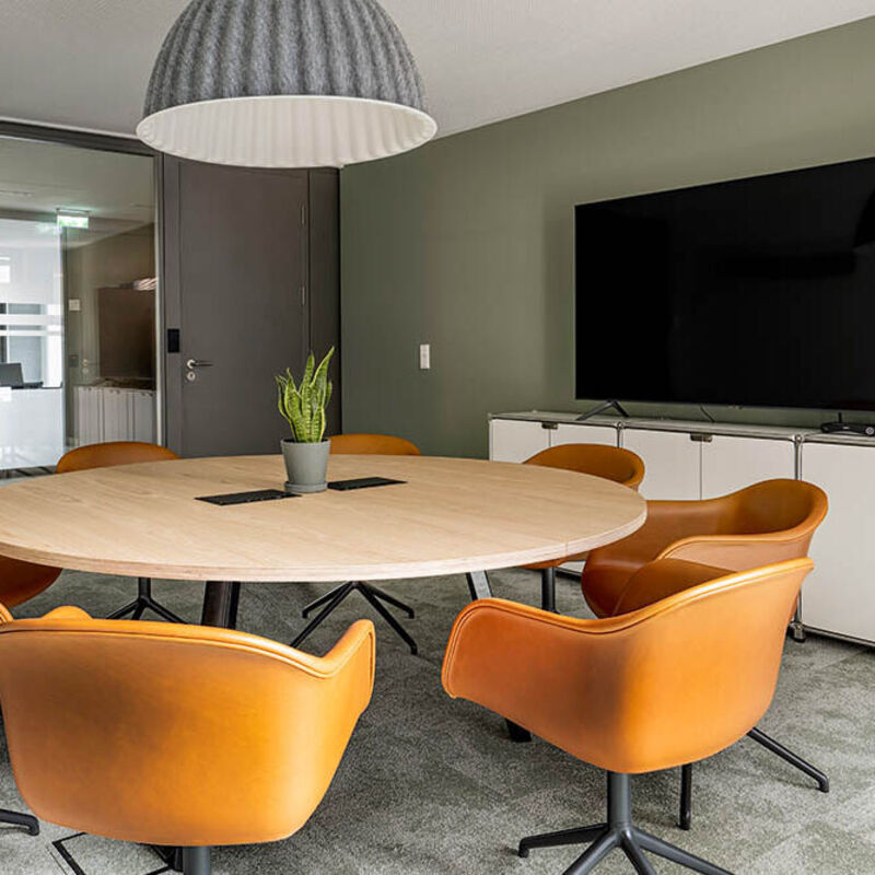 Meetingraum mit rundem Tisch, orangefarbenen Stühlen und Bildschirm vor grüner Wand im Office Club München