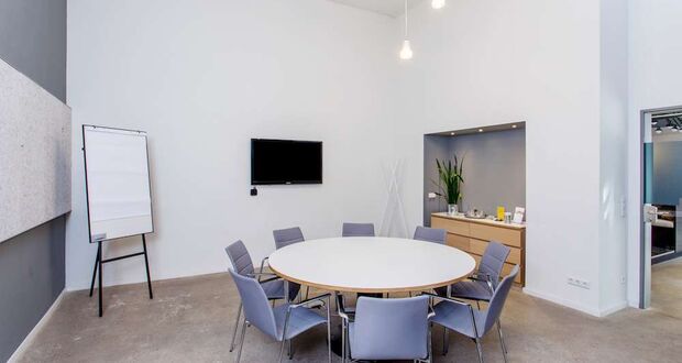 Besprechungsraum mit rundem Tisch, lilafarbenen Stühlen, Bildschirm und Flipchart