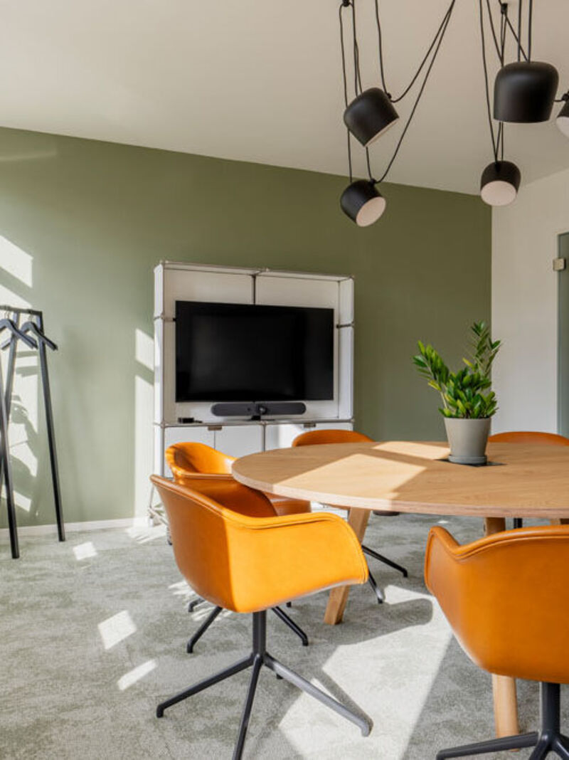 Besprechungsraum mit rundem Holztisch, orangefarbenen Stühlen und Bildschirm im Office Club Köln Hohenzollernring