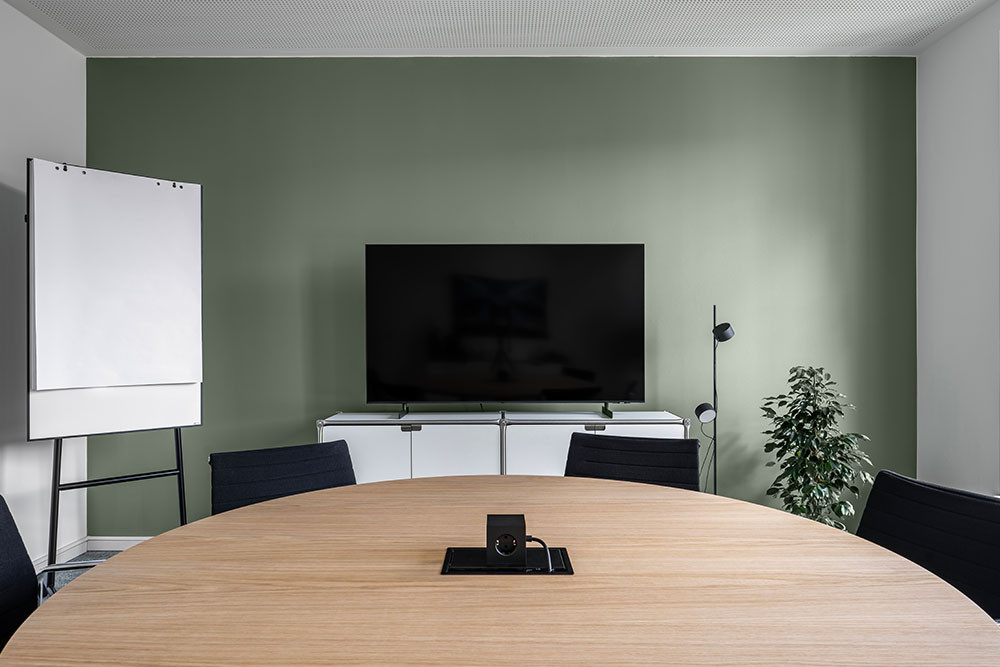 Konferenzraum mit rundem Holztisch, Bildschirm und Flipchart vor grüner Wand im Office Club Hamburg