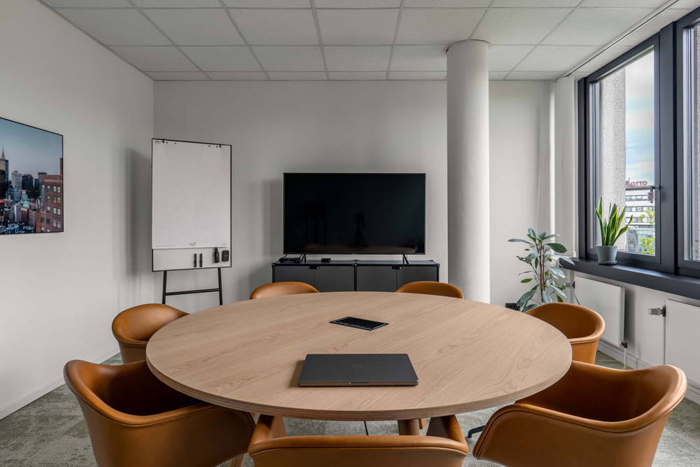 Besprechungsraum mit rundem Holztisch, cognacfarbenen Stühlen, Bildschirm und Flipchart im Office Club Hannover
