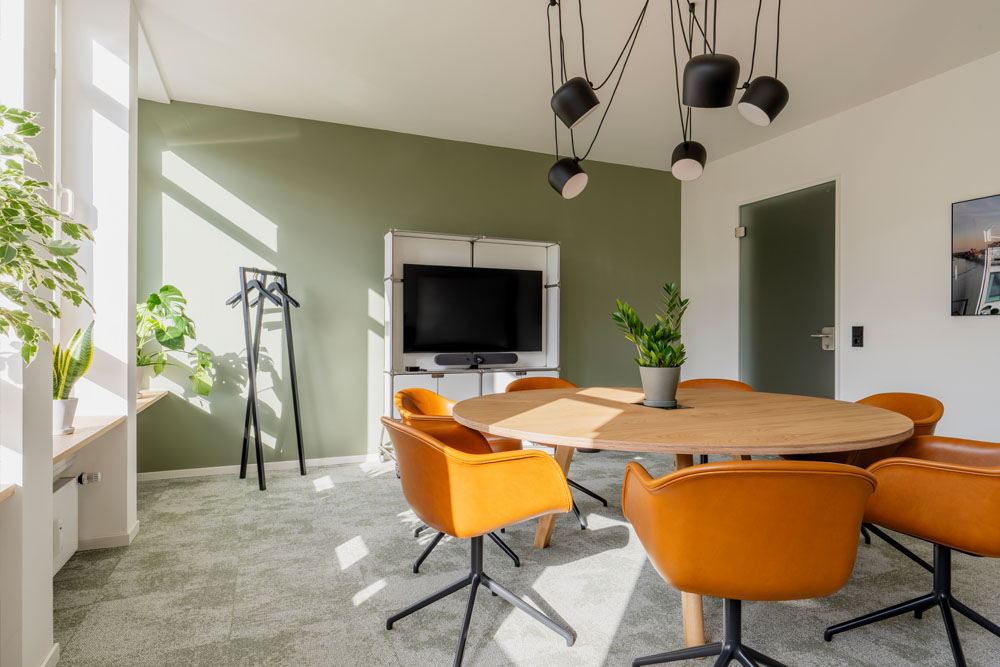 Besprechungsraum mit rundem Holztisch, orangefarbenen Stühlen und Bildschirm im Office Club Köln Hohenzollernring