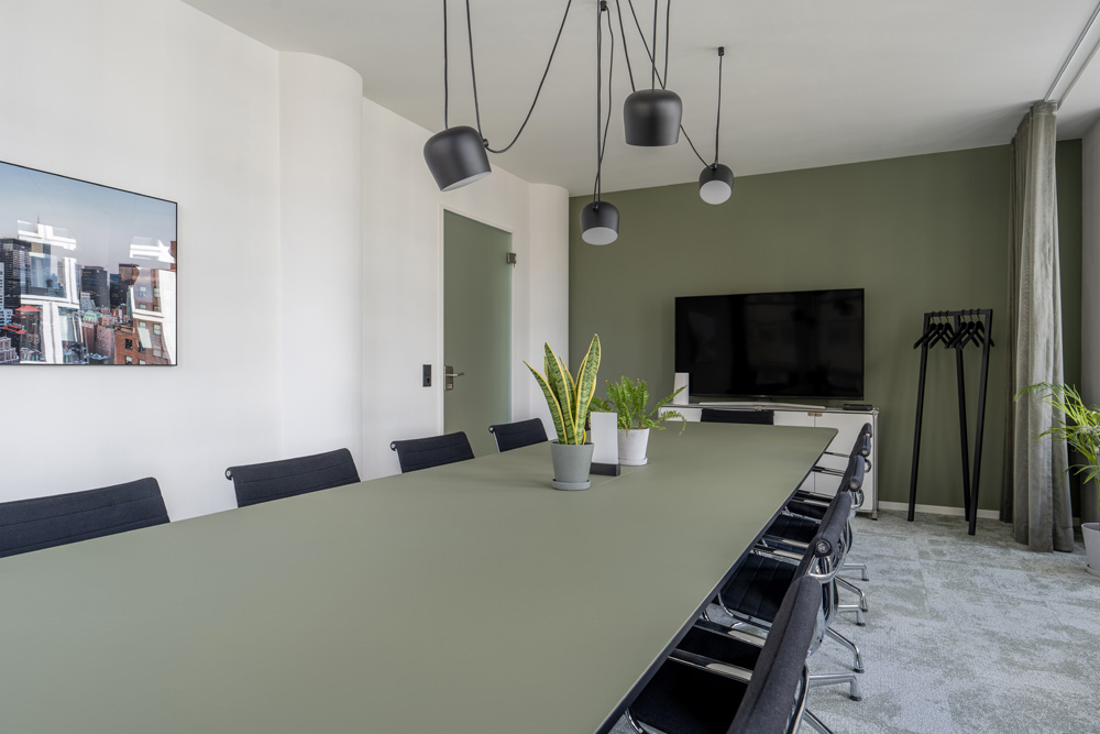 Seminarraum mit langem Tisch, schwarzen Stühlen und Bildschirm vor grüner Wand im Office Club Köln