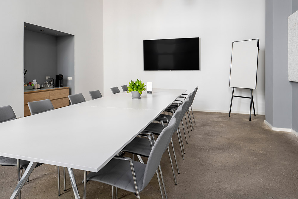 Konferenzraum mit langem weißem Tisch, Bildschirm, Flipchart und Kaffeeecke im Office Club Berlin
