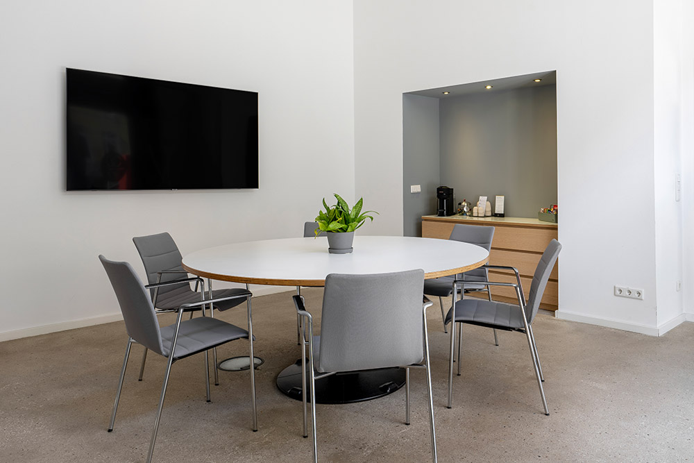 Konferenzraum mit rundem weißem Tisch, grauen Stühlen, Bildschirm und Kaffeeecke im Office Club Berlin