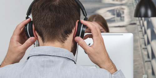 Mann setzt sich am Schreibtisch Kopfhörer auf, im Hintergrund eine Kollegin